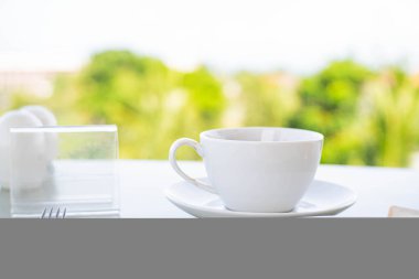 Sehpanın üzerine koyulmuş yeşil arka planı bulanık beyaz bir kahve fincanı. Bir dilim ekmek ve reçel ile bir tabak ön planda kısmen görünür..