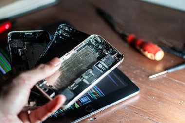 Parçalanmış akıllı telefon parçalarını ekranda kodlanmış bir tabletin üzerinde tutan biri. Aletler ve elektronik parçalar ahşap masaya saçılmış..