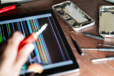 Tahta bir masada elektronik aletleri tamir eden biri. Resim, renkli çizgileri, parçalanmış akıllı telefonları ve çeşitli tamir araçlarını gösteren hasarlı ekranlı bir tableti gösterir.