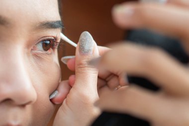 Gözüne makyaj yapmak ya da temizlemek için pamuk çubuğu kullanan birini yakından çek. Parlak bir ojesi var ve elinde bir ayna tutuyor..