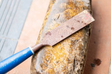 Paslı bir keskinin yakın çekimi yıpranmış bir bilenme taşına yerleştirilmiş. Keskinin mavi bir sapı var ve aşınma ve pas belirtileri gösteriyor. Keskinleştirme taşı eski ve pürüzlü bir dokuya sahip gibi görünüyor..