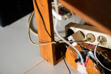 Tahta bir masanın altında birden fazla fiş ve kablo bulunan dağınık bir güç şeridi. Kablolar birbirine dolanmış ve yakınlarda bir karton kutu ile bölge düzensiz görünüyor..