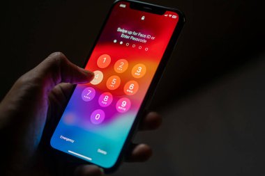 Şifre ekranı görüntülenmiş akıllı bir telefon tutan bir kişi, bir şifre girilmesi veya aygıtın kilidini açmak için Face ID kullanılması gerektiğini belirtir.
