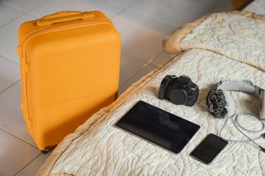 Yorgan kaplı yatağın yanında sarı bir bavul. Yatağın üzerinde bir tablet, akıllı bir telefon, bir kamera ve bir çift kulaklık var..