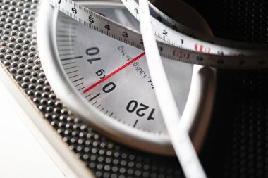 Banyo terazisinin üzerine bir mezura sarılı olan yakın çekimi, ağırlık ölçümü ve sağlık denetimini gösteriyor.