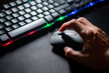 Renkli arka ışıklandırması olan bir klavyenin yanında bilgisayar faresi kullanan birinin elinin yakın plan görüntüsü..