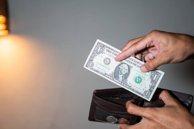 Bir dolarlık banknot ve içinde bozuk paralar olan kahverengi bir cüzdan tutan biri. Arka plan sade ve soldaki ışık kaynağı tarafından hafifçe aydınlatılmış..