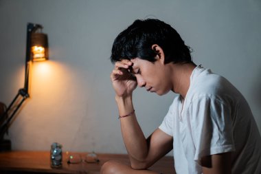 Eli alnında bir masada oturan genç bir adam, stresli ya da derin düşüncelere dalmış görünüyor. Masanın üzerinde bir lamba ve bir gözlük var..