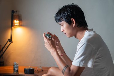 Beyaz tişörtlü genç bir adam loş bir odada küçük bir elektronik cihaz üzerinde çalışıyor. Elinde bir devre kartı var ve görünüşe göre onu onarmaya ya da monte etmeye odaklanmış. Bir masa lambası arka planda ışık sağlar.