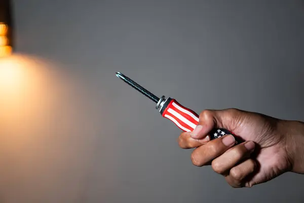 Sapında Amerikan bayrağı tasarımı olan bir tornavidayı arka planda tutan bir el..
