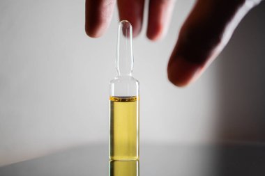 Yansıtıcı bir yüzeye yerleştirilmiş sarı sıvıyla dolu küçük bir cam şişeye uzanan bir el..