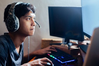 Genç adam oyun oynarken bilgisayar ekranına odaklanmış, kulaklık takıyor ve mikrofon takıyor..