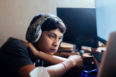 Kulaklık takan genç bir adam bir masada oturuyor, dikkatle bilgisayar ekranına bakıyor. Odaklanmış ve yaptığı şeyle meşgul görünüyor. Ayarlar bir ev ya da ofis ortamını işaret ediyor.