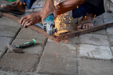 Etrafında kıvılcımlar uçuşan metal bir parçayı kesmek için açı öğütücü kullanan bir işçi. İşçi fayanslı zeminde diz çöküyor ve günlük giysiler giyiyor. Arka planda çeşitli araçlar ve ekipmanlar görünür.