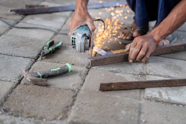 Asfaltlı bir yüzeydeki metal parmaklıkları kıvılcımlar saçarak kesen biri. Pense ve çekiç gibi aletler yakınlarda..