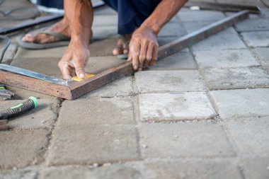 Asfaltlı yüzeyde cetvelle metal çerçeve ölçen biri. Kişi yalınayak ve dışarıda çalışıyor gibi görünüyor..