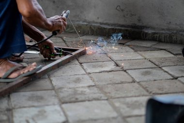 Asfaltlı yüzeyde cetvelle metal çerçeve ölçen biri. Kişi yalınayak ve dışarıda çalışıyor gibi görünüyor..