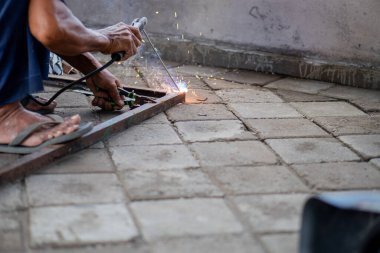 Asfaltlı yüzeyde cetvelle metal çerçeve ölçen biri. Kişi yalınayak ve dışarıda çalışıyor gibi görünüyor..