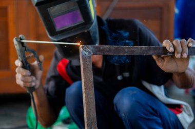 Koruyucu kask takan bir kaynakçı, metal bir çerçeve kaynatıyor, kıvılcımlar ve dumanlar görünür..