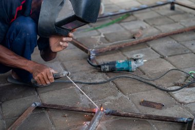 Kaynak miğferi takan bir adam, metal parçaları asfalt yüzeyde birbirine kaynaştırıyor. Yanında elektrikli bir alet var..