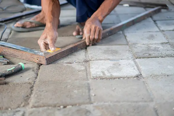 Asfaltlı yüzeyde cetvelle metal çerçeve ölçen biri. Kişi yalınayak ve dışarıda çalışıyor gibi görünüyor..