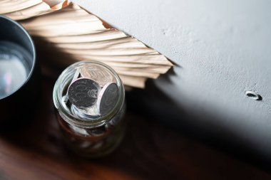 Tahta bir yüzeye yerleştirilmiş bozuk paralarla dolu cam bir kavanoz. Yanında da bir yığın kağıt ve siyah bir kap var..