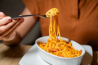Beyaz bir tabaktan çatalla spagetti yiyen birinin yakın çekimi. Spagetti kremalı portakal sosuyla kaplı ve küçük et parçalarıyla süslenmiş..