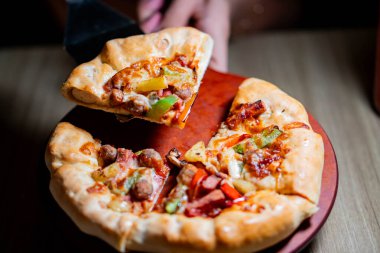 Kalın hamurlu taze pişmiş pizza. Üstünde dolma biber, sosis ve peynir gibi çeşitli malzemeler var. Tahta bir kürek üzerinde servis ediliyor..