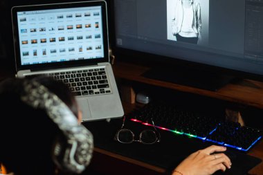 Uzun saçlı, beyaz tişörtlü ve boynunda büyük kulaklıklar olan bir kadın elinde fotoğraf düzenleme yazılımı olan bir bilgisayarın önünde oturuyor..