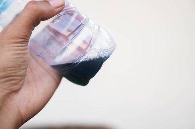 İçinde az miktarda koyu sıvı bulunan plastik bir şişeyi tutan bir elin yakın çekimi. Arka plan sade ve bulanık..