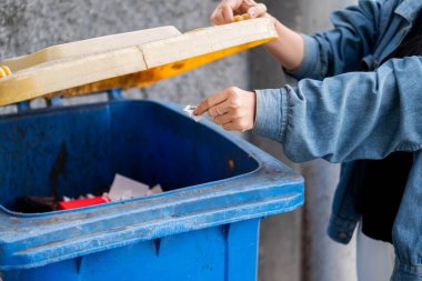 Kot ceketli bir insan sarı kapaklı mavi bir geri dönüşüm kutusuna kağıt atıyor..