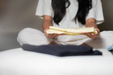 Yatakta bağdaş kurmuş, giysilerini katlayan biri. Ellere ve kıyafetlere odaklanır, kişinin yüzü görünmezdir..