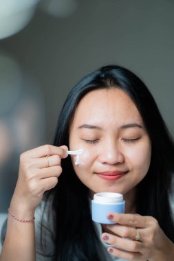 Küçük bir spatulayla yüz kremi süren ve gözleri kapalı gülümseyen bir kadın. O, sol elinde bir krem kavanozu tutuyor ve kremi yüzüne sürmek için sağ elini kullanıyor. Arkaplan bulanık.