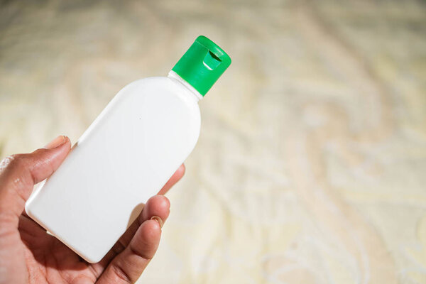 Рука, держащая маленькую белую пластиковую бутылку с зеленой крышкой, на размытом фоне.