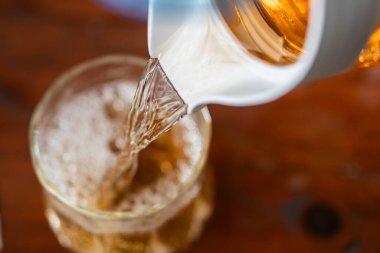 Beyaz bir sürahiden bardağa dökülen bir içeceğin yakından çekimi. İçkinin baloncuklu kahverengi bir rengi var gibi görünüyor. Bu da karbonatlı içecek ya da buzlu çay olabileceğini gösteriyor. Ahşap masa yüzeyi sahneye sıcaklık katıyor..