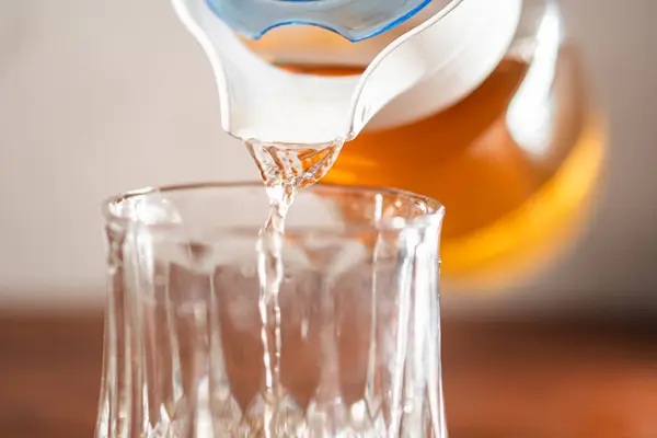 stock image A close-up shot of a glass being filled with a golden liquid from a pitcher. The liquid appears to be tea or a similar beverage, with a smooth pouring action captured. The background is softly blurred, emphasizing the pouring process.