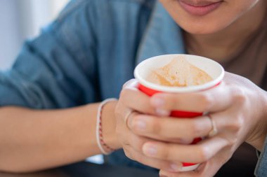 Elinde kırmızı bir kahve fincanı olan ve elinde köpüklü bir içecek olan birinin yakın çekimi. Arka plan hafifçe bulanık, sıcak içeceği vurguluyor..