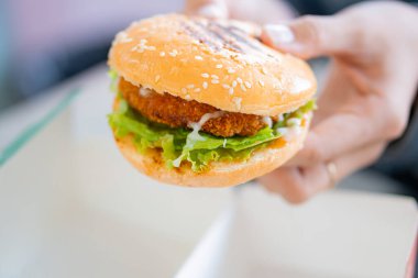 Kızarmış tavuk, taze marul ve mayonez ile dolu susamlı çörek ekmeği tutan yakın plan bir el, bulanık bir arka plan üzerine kurulmuş..