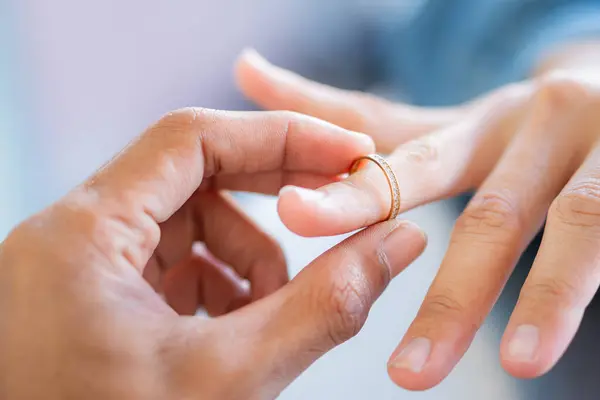 stock image A close-up of a hand holding a gold ring, with another hand in the background. The focus is on the ring, symbolizing love or commitment.