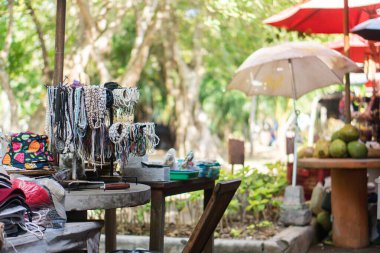 Ahşap bir masanın üzerinde renkli mücevherlerin sergilendiği canlı bir açık hava piyasa sahnesi. Arka planda, yemyeşil ve şemsiyeler gölge oluştururken, çeşitli eşyalar etrafa dağılıp canlı bir atmosfer yaratıyorlar..