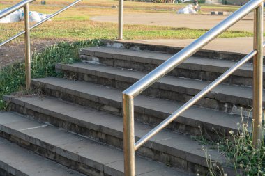 Paslanmaz çelik parmaklıklı beton merdiven setinin yakın çekim görüntüsü. Merdivenler çok yıpranmış, kullanım belirtileri gösteriyor ve etrafı ot ve yabani otlarla çevrili. Arka planda çöp torbaları ve açık bir alan var..