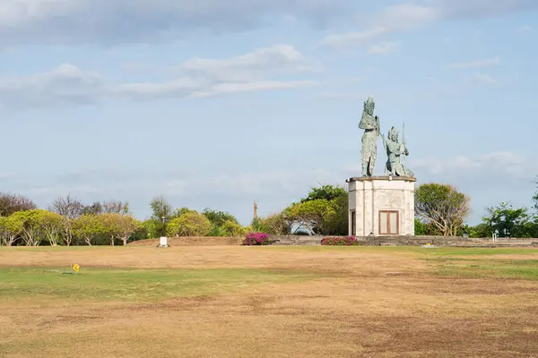 stock image A large statue of two figures stands on a grassy area with sparse vegetation. The sky is partly cloudy, and there are some trees and shrubs in the background. The ground appears dry and brown, indicating a lack of water.