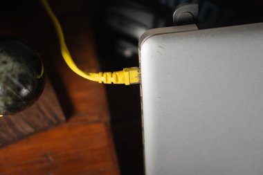 Gümüş bir dizüstü bilgisayara takılmış sarı bir ethernet kablosunun yakından görüntüsü. Arkaplanda ahşap bir yüzey ve dekoratif bir nesne var..