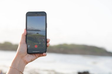 Manzaralı bir okyanus manzarası önünde akıllı bir telefon tutan kişi manzarayı yakalıyor. Telefon ekranı aynı görüntüyü gösterir, dalgaları ve açık gökyüzünü gösterir.
