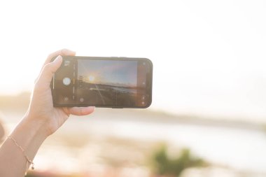 Günbatımını kaydetmek için elinde akıllı bir telefon tutan biri, parlak bir güneş ve manzaralı bir arka plan. Telefon ekranı kayıt arayüzünü gösterir.