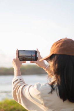 Akıllı telefonuyla manzaralı bir görüntü yakalayan bir kadın, açık renk gömlek ve şapka takıyor. Arkaplanda açık bir gökyüzü ve uzak bir manzara var..