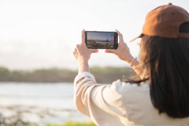 Akıllı telefonuyla manzaralı bir görüntü yakalayan bir kadın, açık renk gömlek ve şapka takıyor. Arkaplanda açık bir gökyüzü ve uzak bir manzara var..