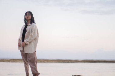 Yeşil deniz yosunlarıyla kaplı kayalık bir sahilde yalınayak duran bir insan gün batımında sığ sulara yansıyor. Arka planda sakin bir deniz ve pastel bir gökyüzü var..