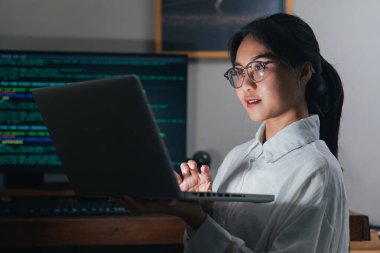 Gözlüklü genç bir kadın loş bir odada dizüstü bilgisayar üzerinde çalışıyor. Odaklanmış ve meşgul görünüyor. Arka planda bilgisayar ekranı görüntüleme kodu var..