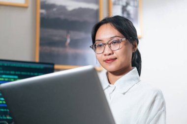 Modern ofis ortamında dizüstü bilgisayarda çalışan gözlüklü genç bir kadın. Çerçeveli fotoğraflar ve kod gösteren bir bilgisayar ekranıyla odaklanmış ve meşgul görünüyor..